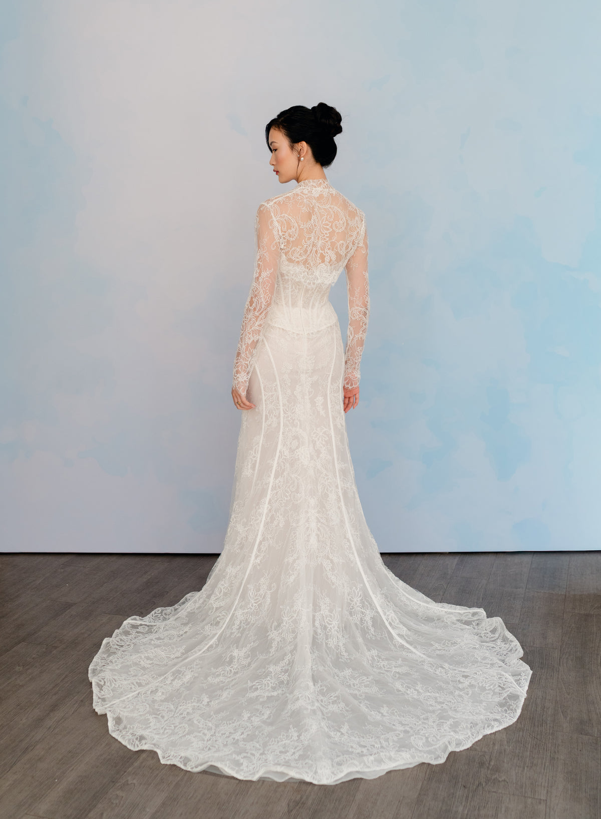 A woman wearing a long-sleeve lace wedding gown is shown from the back, standing on a dark wood floor against a light blue studio backdrop. / Slim lace gown with semi-sheer bodice with sweetheart neckline and bustier cut. Small peplums on sides hips. Invisible zipper back closure. Matching lace bolero with long sleeves.