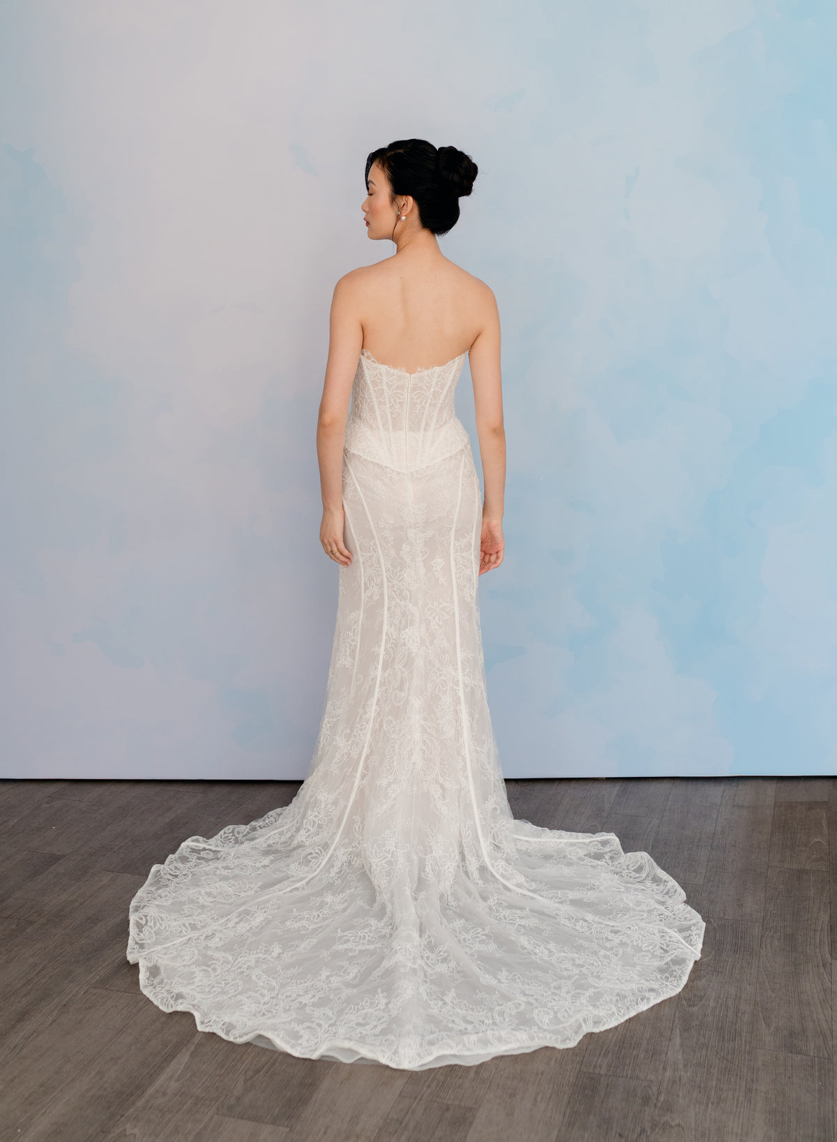 A woman stands with her back to the camera wearing a strapless lace wedding gown with a fitted bodice and a long train, photographed against a soft blue studio backdrop on a wood floor. / Slim lace gown with semi-sheer bodice with sweetheart neckline and bustier cut. Small peplums on sides hips. Invisible zipper back closure. Matching lace bolero with long sleeves.