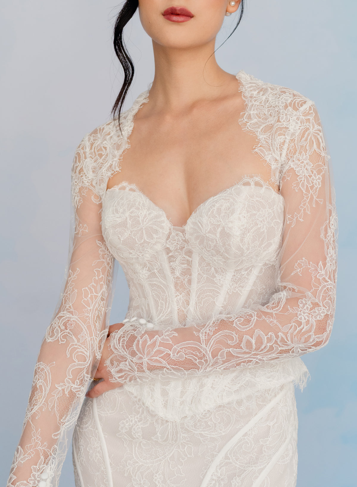 A close-up of a model wearing a white lace bridal gown with long sheer lace sleeves and a sweetheart neckline, set against a soft light blue background. / Slim lace gown with semi-sheer bodice with sweetheart neckline and bustier cut. Small peplums on sides hips. Invisible zipper back closure. Matching lace bolero with long sleeves.