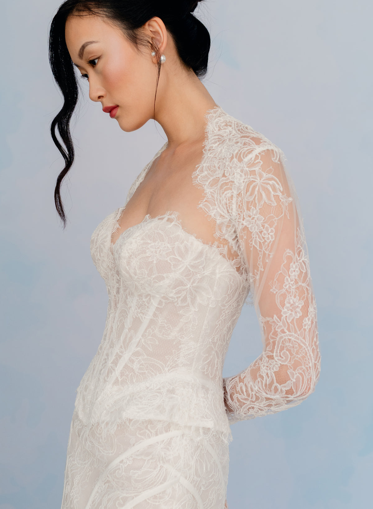 A woman wearing a white lace bridal gown with long sheer sleeves and a sweetheart neckline, photographed against a soft light blue studio background. / Slim lace gown with semi-sheer bodice with sweetheart neckline and bustier cut. Small peplums on sides hips. Invisible zipper back closure. Matching lace bolero with long sleeves.