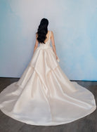 A woman viewed from behind wearing a voluminous ivory satin wedding gown with a long train, standing against a light blue wall on a dark wood floor. / Dramatic, sculpted Mikado gown with lace placement.