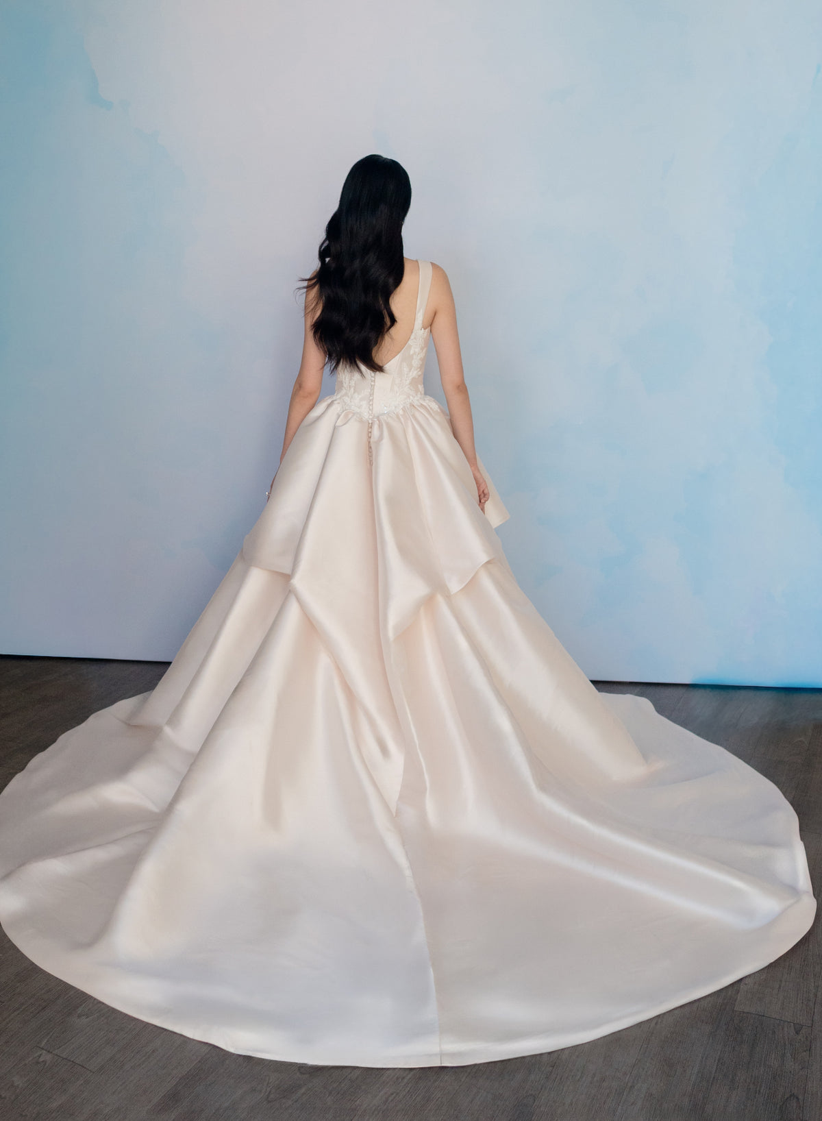 A woman viewed from behind wearing a voluminous ivory satin wedding gown with a long train, standing against a light blue wall on a dark wood floor. / Dramatic, sculpted Mikado gown with lace placement.