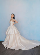 A woman modeling a voluminous champagne-colored satin ball gown with a long train, standing against a textured light blue wall. / Dramatic, sculpted Mikado gown with lace placement.