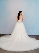 A woman wearing a strapless white wedding gown with a fitted lace-up corset back and a full tulle train, photographed from behind against a soft blue studio backdrop. / Semi sheer Basque waist bodice with lace appliques and dramatic tulle skirt.