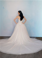 A woman stands with her back to the camera wearing a strapless white wedding gown with a fitted lace corset-style bodice and a very full tulle train on a gray floor against a pale blue studio backdrop. / Semi sheer Basque waist bodice with lace appliques and dramatic tulle skirt.