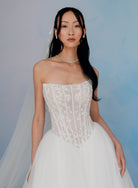A woman modeling a strapless white lace wedding gown with a fitted lace bodice and a full tulle skirt, posed against a soft blue studio backdrop. / Semi sheer Basque waist bodice with lace appliques and dramatic tulle skirt.