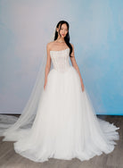 A woman modeling a strapless white wedding gown with a structured lace corset bodice and a full tulle skirt, standing against a soft blue studio backdrop. / Semi sheer Basque waist bodice with lace appliques and dramatic tulle skirt.