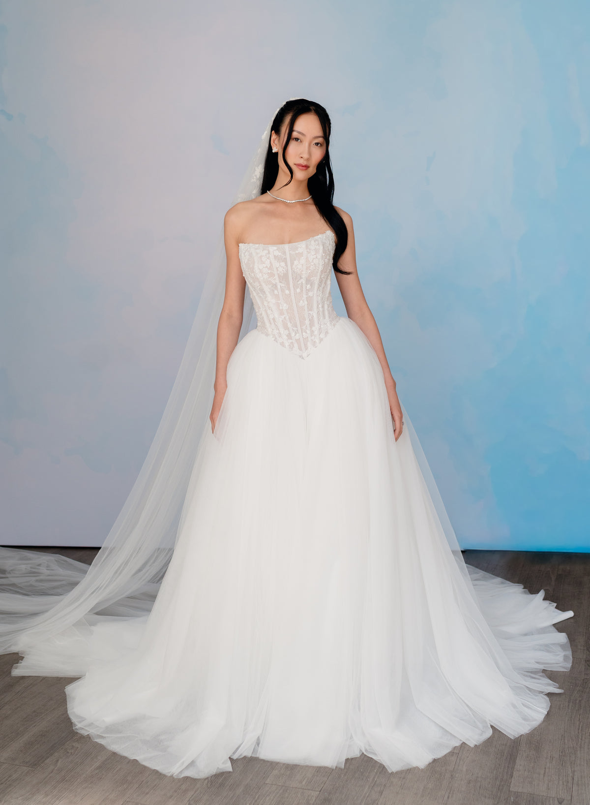A woman modeling a strapless white wedding gown with a structured lace corset bodice and a full tulle skirt, standing against a soft blue studio backdrop. / Semi sheer Basque waist bodice with lace appliques and dramatic tulle skirt.