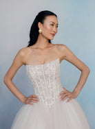 A woman modeling a strapless white lace corset dress with a voluminous tulle skirt, posed against a soft light blue studio backdrop. / Semi sheer Basque waist bodice with lace appliques and dramatic tulle skirt.