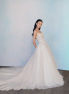 A woman modeling a strapless white wedding gown with a fitted embellished bodice and a voluminous tulle skirt with a long train, standing against a soft light blue studio backdrop. / Semi sheer Basque waist bodice with lace appliques and dramatic tulle skirt.
