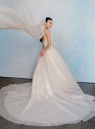 A woman modeling a strapless ivory wedding gown with a voluminous tulle skirt and matching sheer veil, posed against a soft light blue studio backdrop. / Semi sheer Basque waist bodice with lace appliques and dramatic tulle skirt. / Veil with lace.