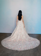 A woman stands with her back to the camera wearing a sleeveless, floor-length lace gown with a very wide train on a dark wood floor against a pale blue wall. / All over multicolored lace A-line gown.