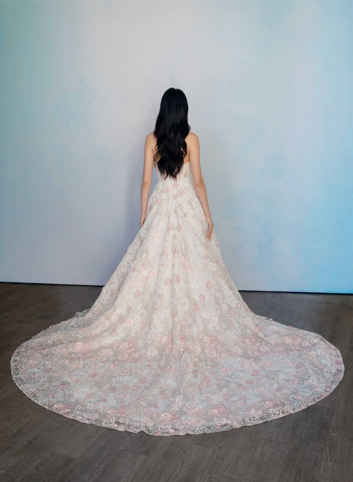 A woman stands with her back to the camera wearing a sleeveless, floor-length lace gown with a very wide train on a dark wood floor against a pale blue wall. / All over multicolored lace A-line gown.