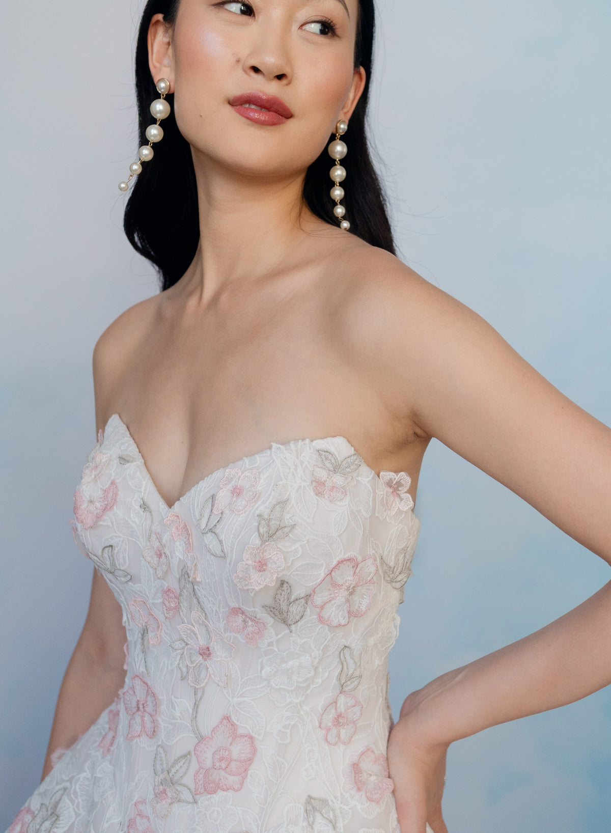 A woman wearing a strapless white lace dress with pink floral embroidery and long pearl drop earrings, posed against a soft light blue studio background. / All over multicolored lace A-line gown.