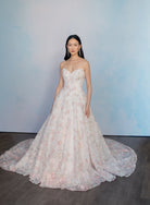 A woman stands in a strapless ball gown with a sweetheart neckline, featuring intricate floral lace embroidery in pale pink and white over a sheer tulle base. The dress has a full skirt with a long train, set against a soft blue watercolor-style backdrop on a dark wood floor. / All over multicolored lace A-line gown.