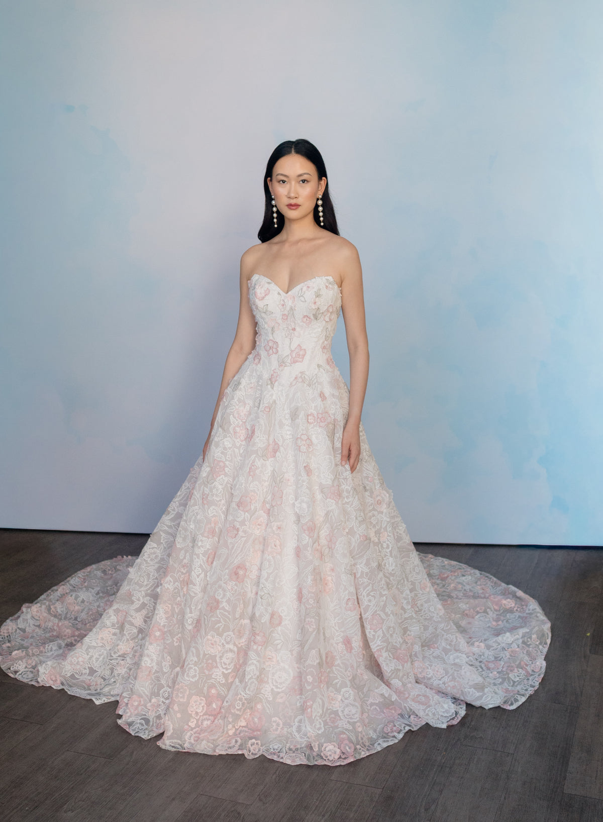 A woman stands in a strapless ball gown with a sweetheart neckline, featuring intricate floral lace embroidery in pale pink and white over a sheer tulle base. The dress has a full skirt with a long train, set against a soft blue watercolor-style backdrop on a dark wood floor. / All over multicolored lace A-line gown.