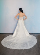 A woman stands with her back to the camera wearing a strapless white wedding gown with a fitted lace bodice and a long sheer overskirt with a wide lace hem, posed against a light blue wall on a dark wood floor. / Slim mikado style with pleated bodice with straight neckline and zipper/buttons back closure. Lace scallop decoration on bodice and on skirt sides. Matching mikado detachable train with sheer back area covered with lace.
