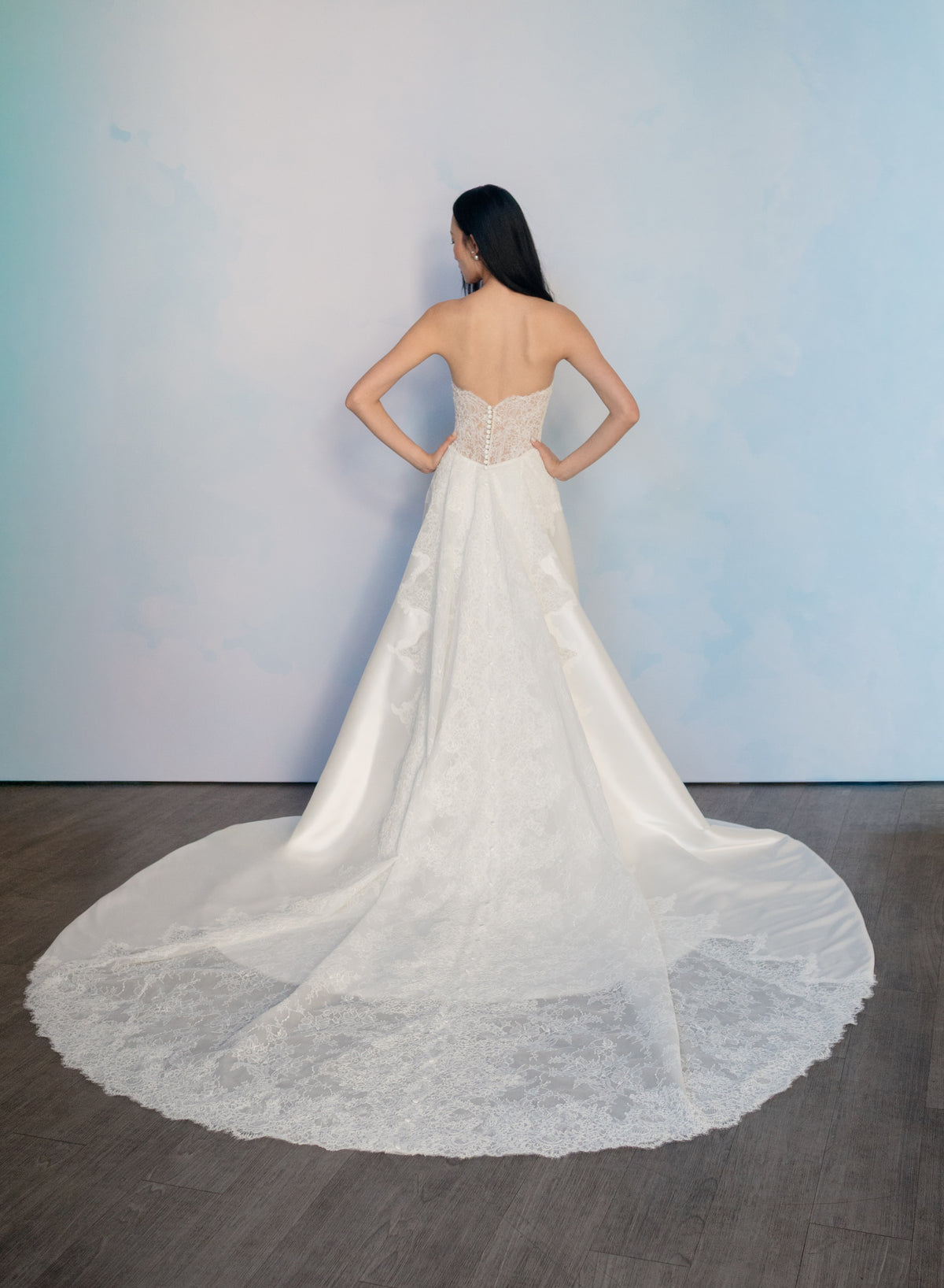 A woman stands with her back to the camera wearing a strapless white wedding gown with a fitted lace bodice and a long sheer overskirt with a wide lace hem, posed against a light blue wall on a dark wood floor. / Slim mikado style with pleated bodice with straight neckline and zipper/buttons back closure. Lace scallop decoration on bodice and on skirt sides. Matching mikado detachable train with sheer back area covered with lace.