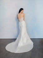 A woman wearing a strapless white wedding gown with a fitted lace bodice and a long satin train, photographed from the back against a pale blue wall and dark wood floor. / Slim mikado style with pleated bodice with straight neckline and zipper/buttons back closure. Lace scallop decoration on bodice and on skirt sides. Matching mikado detachable train with sheer back area covered with lace.