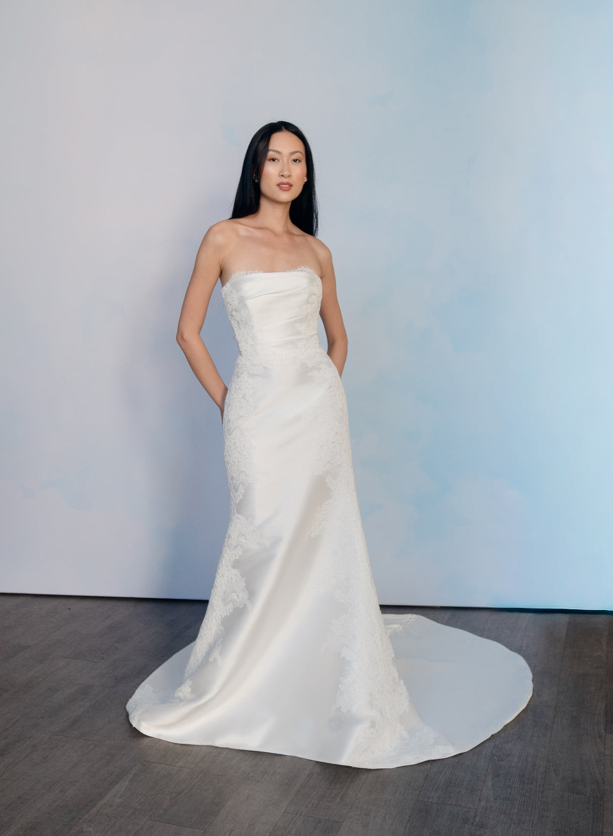 A woman modeling a strapless white wedding gown with a fitted bodice, subtle lace or embroidered texture, and a slight train, standing against a pale blue studio backdrop on a dark wood floor. / Slim mikado style with pleated bodice with straight neckline and zipper/buttons back closure. Lace scallop decoration on bodice and on skirt sides. Matching mikado detachable train with sheer back area covered with lace.