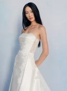 A woman modeling a strapless white wedding gown with lace detailing and a satin skirt, posed against a soft light blue studio background. / Slim mikado style with pleated bodice with straight neckline and zipper/buttons back closure. Lace scallop decoration on bodice and on skirt sides. Matching mikado detachable train with sheer back area covered with lace.