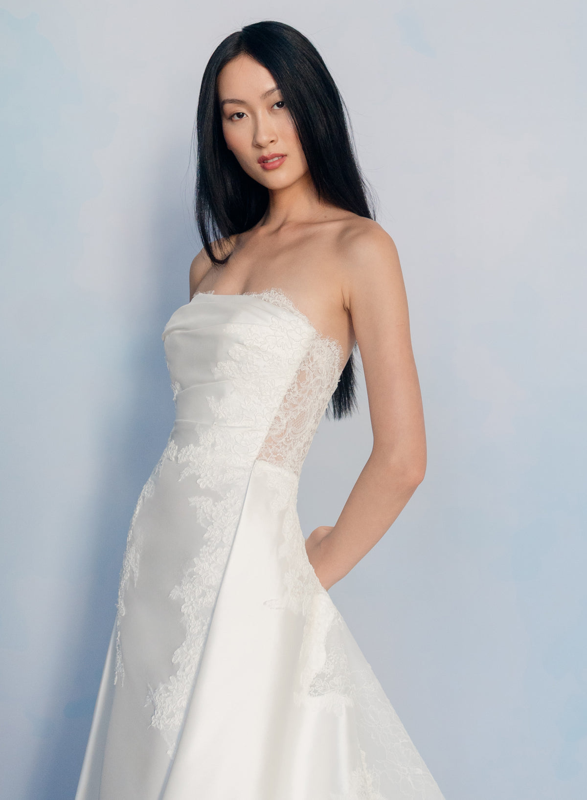 A woman modeling a strapless white wedding gown with lace detailing and a satin skirt, posed against a soft light blue studio background. / Slim mikado style with pleated bodice with straight neckline and zipper/buttons back closure. Lace scallop decoration on bodice and on skirt sides. Matching mikado detachable train with sheer back area covered with lace.