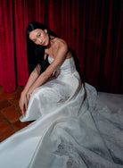 A woman in a strapless white satin and lace gown sits on a red velvet curtain, with terracotta tile flooring visible in the lower left. / Slim mikado style with pleated bodice with straight neckline and zipper/buttons back closure. Lace scallop decoration on bodice and on skirt sides. Matching mikado detachable train with sheer back area covered with lace.