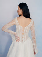 A woman wearing a white lace bridal gown, shown from the back. The dress features long sheer lace sleeves, a deep V-back, and an open lace-up corset-style back. / All over lace gown with long sleeve, basque waist and A-line skirt with lace up back.