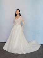 A woman modeling a long-sleeved white lace wedding gown with a deep V-neckline and a full skirt with a train, standing against a light blue textured backdrop. / All over lace gown with long sleeve, basque waist and A-line skirt with lace up back.