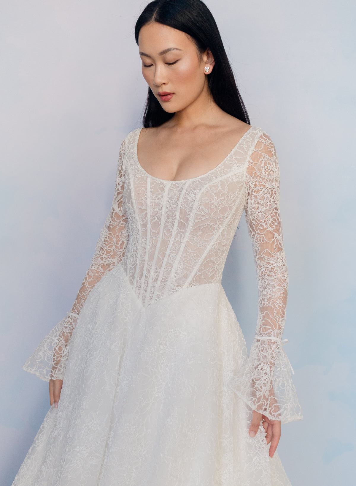 A woman wearing a white lace wedding gown with long sheer sleeves, a scoop neckline, and a full skirt, posed against a soft light blue studio background. / All over lace gown with long sleeve, basque waist and A-line skirt with lace up back.