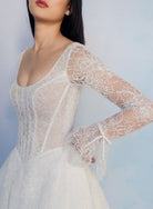 A woman wearing a white lace bridal gown with long sheer lace sleeves and a fitted bodice, photographed against a soft light blue studio background. / All over lace gown with long sleeve, basque waist and A-line skirt with lace up back.