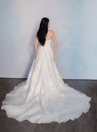 A woman stands with her back to the camera wearing a strapless white wedding gown with a full, flowing train on a dark wood floor against a pale blue wall. / Organza and lace princess ballgown with lace applique details.