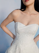A close-up of a model wearing a strapless white wedding dress with a sweetheart neckline, smooth satin-like fabric, and floral lace applique on the bodice, against a soft blue studio background. / Organza and lace princess ballgown with lace applique details.