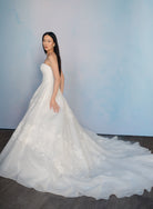 A woman modeling a strapless white wedding gown with a long train, photographed against a soft light blue studio backdrop. / Organza and lace princess ballgown with lace applique details.
