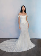 A woman modeling an off-the-shoulder white lace wedding gown with a structured bodice and a long train, standing against a soft blue-gray studio backdrop. / Slim style mock 2 piece with satin bodice and semi-sheer lace skirt. Scooped neckline with off-shoulder straps decorated with delicate lace scallop edge, invisible zipper back closure.