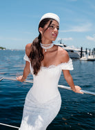 A woman wearing a white off-the-shoulder lace dress and matching white hat stands on a yacht railing with blue water and boats in the background. / Slim style mock 2 piece with satin bodice and semi-sheer lace skirt. Scooped neckline with off-shoulder straps decorated with delicate lace scallop edge, invisible zipper back closure.