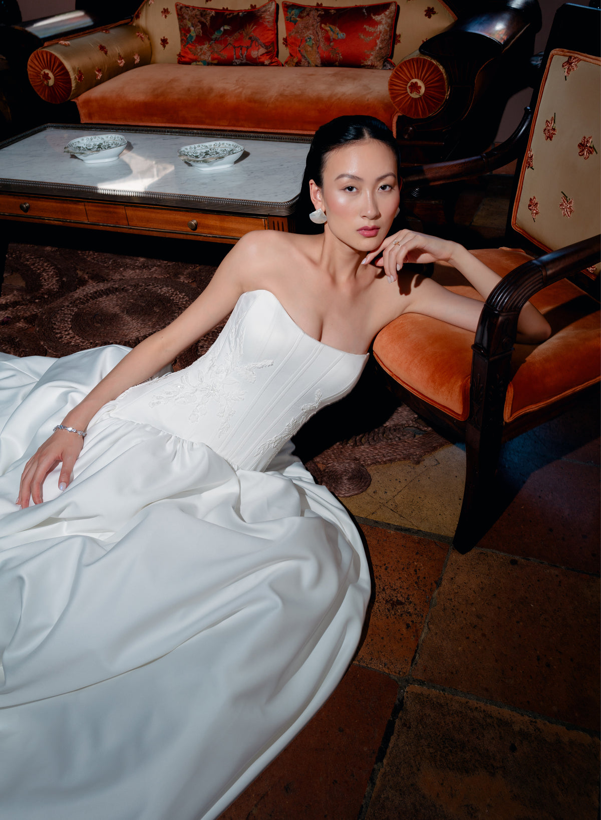 A woman reclining in a luxurious room wearing a strapless white satin gown with a structured bodice and full flowing skirt. She is posed next to an orange velvet armchair with a marble-top coffee table and an ornate sofa in the background. / Dramatic drop waist, double face satin ballgown with placed lace and lace up back detail.