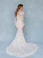 A woman wearing a long-sleeve white lace mermaid-style wedding gown, shown from the back against a soft light blue textured studio backdrop. / All over lace and lace applique slim gown with detachable lining and jacket as well as structured corset.
