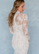 A woman wearing a white lace wedding dress, shown from the back and side against a textured light blue studio backdrop. / All over lace and lace applique slim gown with detachable lining and jacket as well as structured corset.