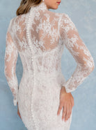 A woman wearing a white lace long-sleeve dress, shown from the back against a light blue textured background. / All over lace and lace applique slim gown with detachable lining and jacket as well as structured corset.