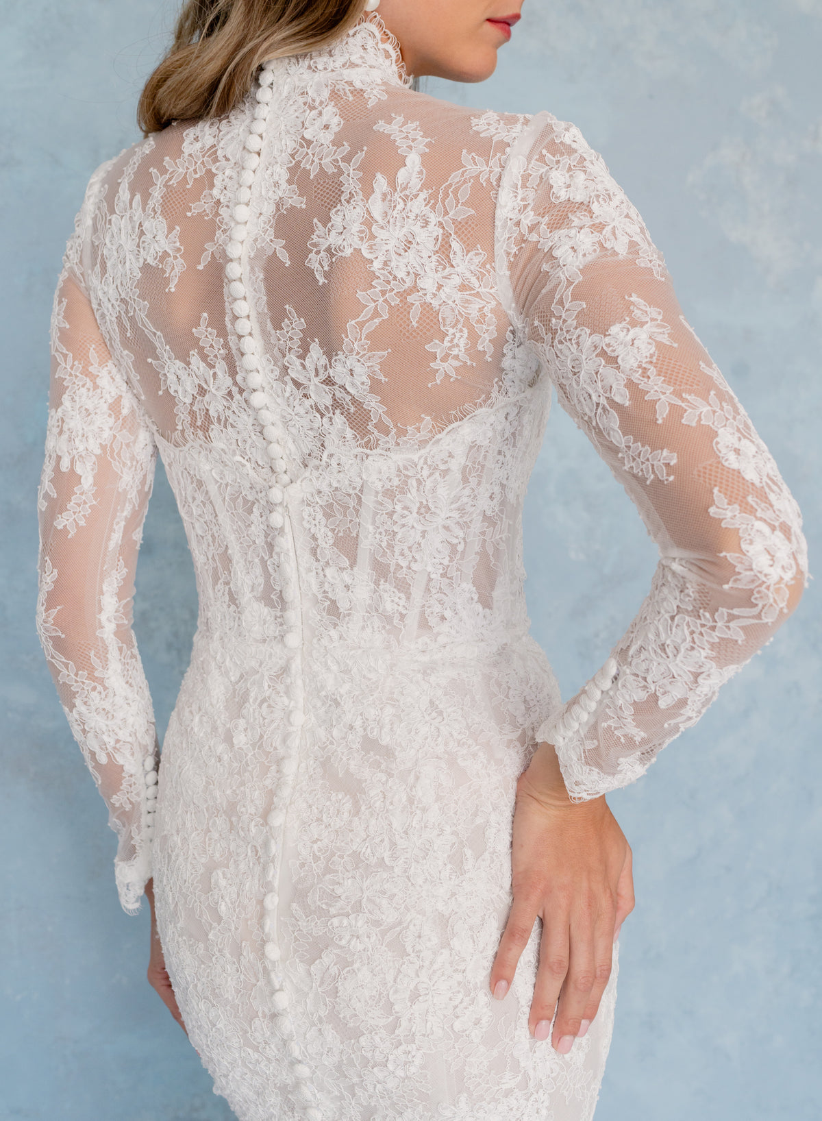 A woman wearing a white lace long-sleeve dress, shown from the back against a light blue textured background. / All over lace and lace applique slim gown with detachable lining and jacket as well as structured corset.