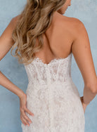 Rear view of a woman wearing a strapless lace wedding dress against a soft blue textured background. / All over lace and lace applique slim gown with detachable lining and jacket as well as structured corset.