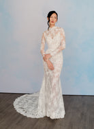 A woman wearing a long-sleeve lace wedding gown with a high mock neck and a train, standing against a light blue backdrop. / All over lace and lace applique slim gown with detachable lining and jacket as well as structured corset.