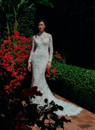 A woman wearing a fitted lace wedding gown with long sleeves and a high neckline stands outdoors on a brick path, surrounded by green hedges and bright red flowers. / All over lace and lace applique slim gown with detachable lining and jacket as well as structured corset.