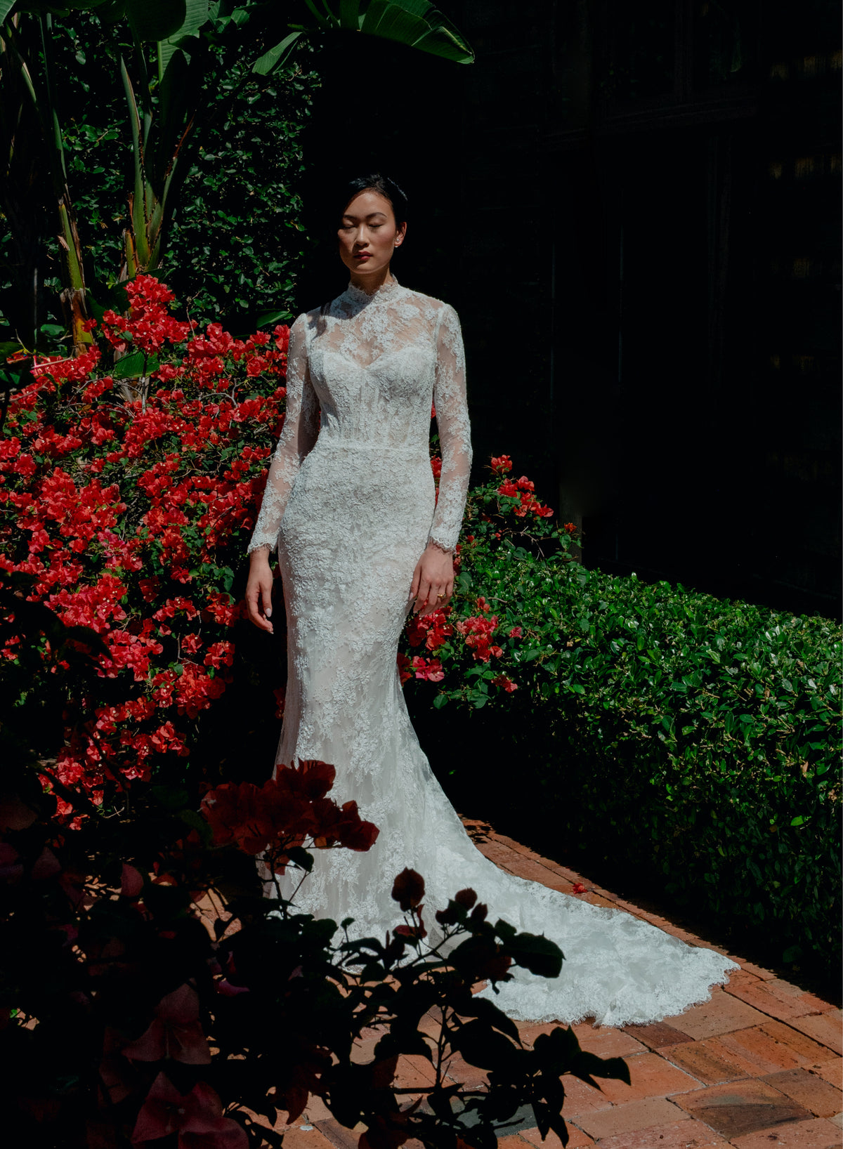 A woman wearing a fitted lace wedding gown with long sleeves and a high neckline stands outdoors on a brick path, surrounded by green hedges and bright red flowers. / All over lace and lace applique slim gown with detachable lining and jacket as well as structured corset.