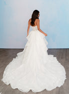 A woman wearing a strapless white wedding gown with a lace corset-style bodice and a full layered tulle train, photographed from the back against a pale blue studio backdrop and wood floor. / All over lace gown with cat eye neckline and soft, dramatic cascading skirt.