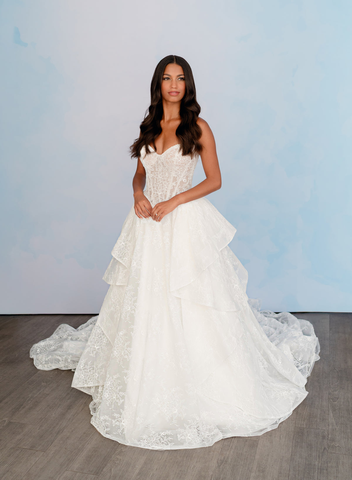 A woman modeling a strapless white lace wedding gown with a sweetheart neckline, fitted bodice, and a full tiered tulle skirt with a long train, standing against a pale blue studio backdrop. / All over lace gown with cat eye neckline and soft, dramatic cascading skirt.