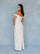 A woman modeling an off-the-shoulder white lace maxi dress with a fitted corset-style bodice and a front slit, standing against a textured light blue studio backdrop. / All over lace column gown with a dramatic matching overskirt.