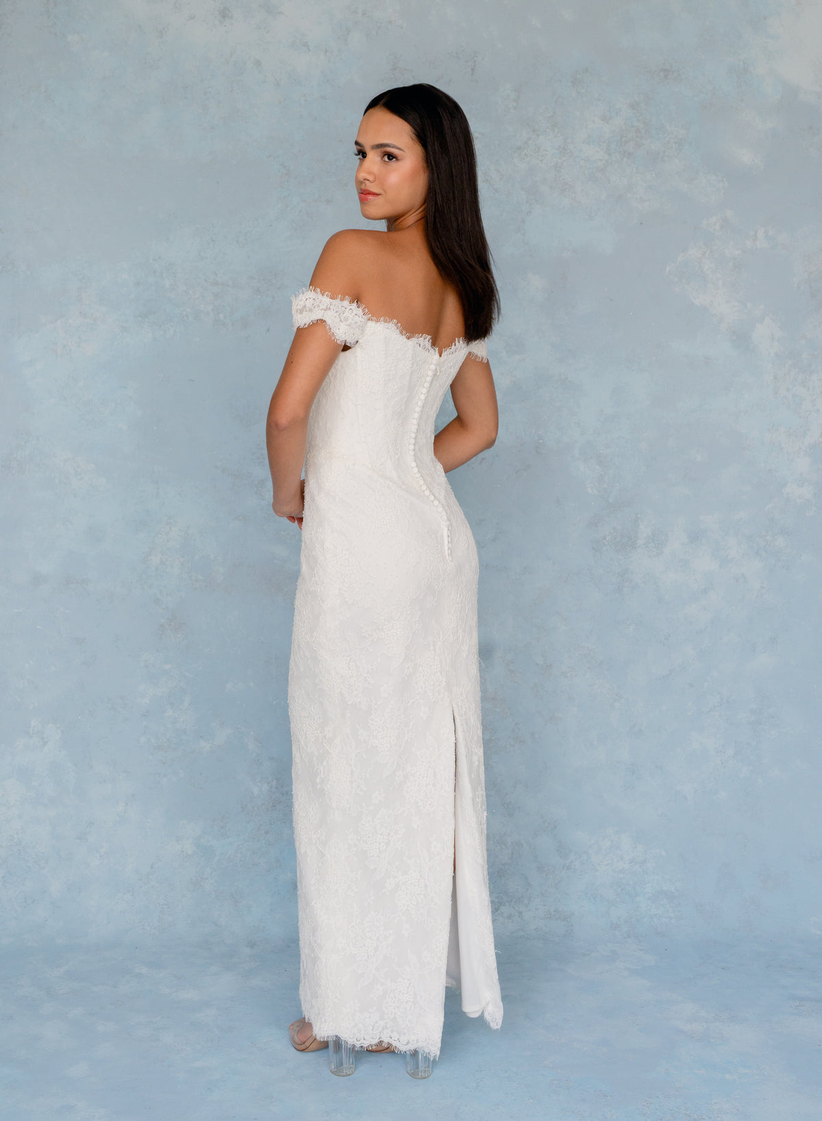 A woman modeling an off-the-shoulder white lace maxi dress with a fitted corset-style bodice and a front slit, standing against a textured light blue studio backdrop. / All over lace column gown with a dramatic matching overskirt.