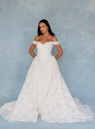 A woman modeling an off-the-shoulder white lace wedding gown with a fitted bodice and full skirt, standing against a textured light blue backdrop. / All over lace column gown with a dramatic matching overskirt.